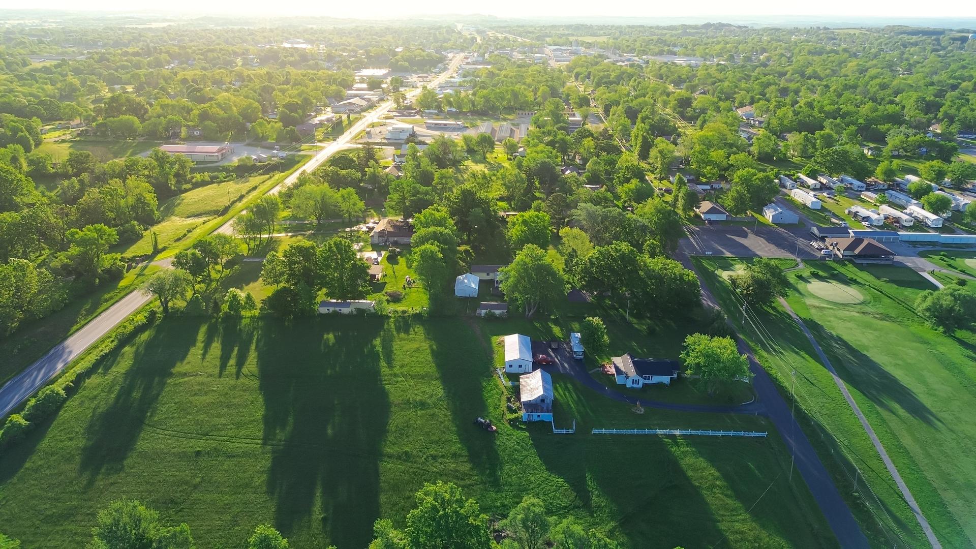 Service road with power line in countryside Mountain Grove Missouri thru lush greenery trees, meadows and farm houses with ponds in agricultural area, peaceful rural lots in Midwest, aerial view